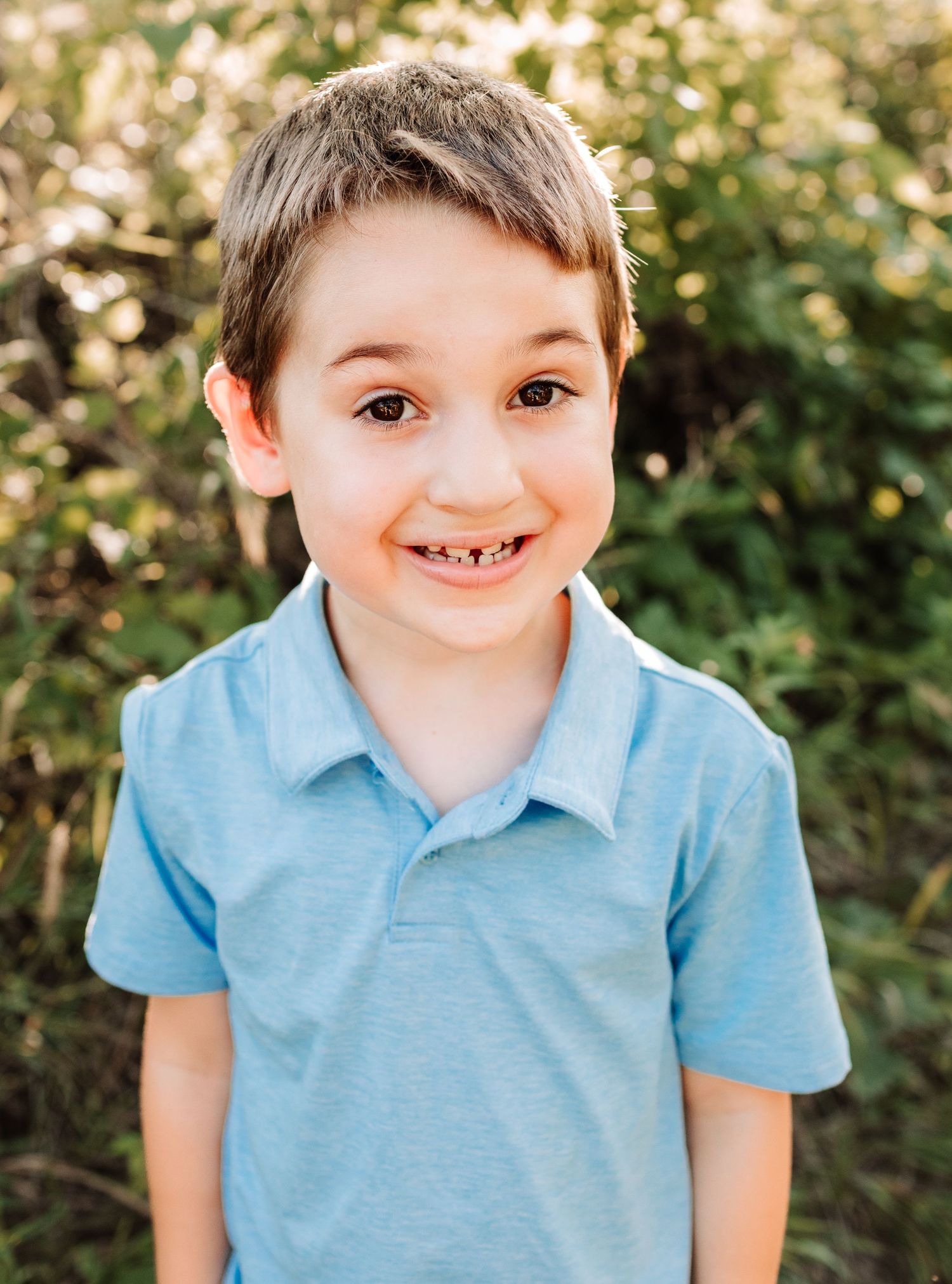 Smiling young boy with missing front teeth wearing a light blue polo shirt outdoors.