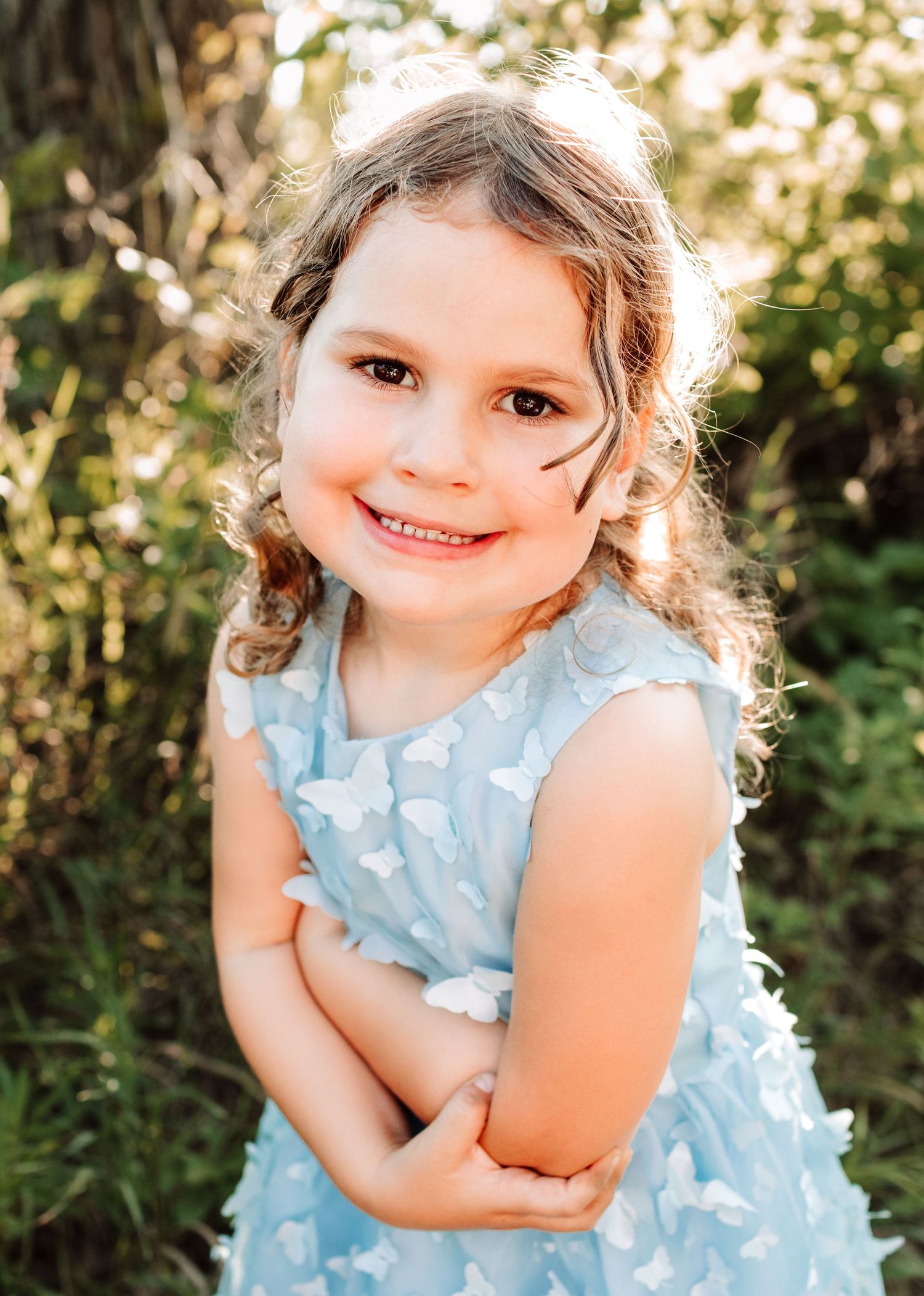 Adorable little girl in a blue butterfly dress smiling with arms crossed outdoors.
