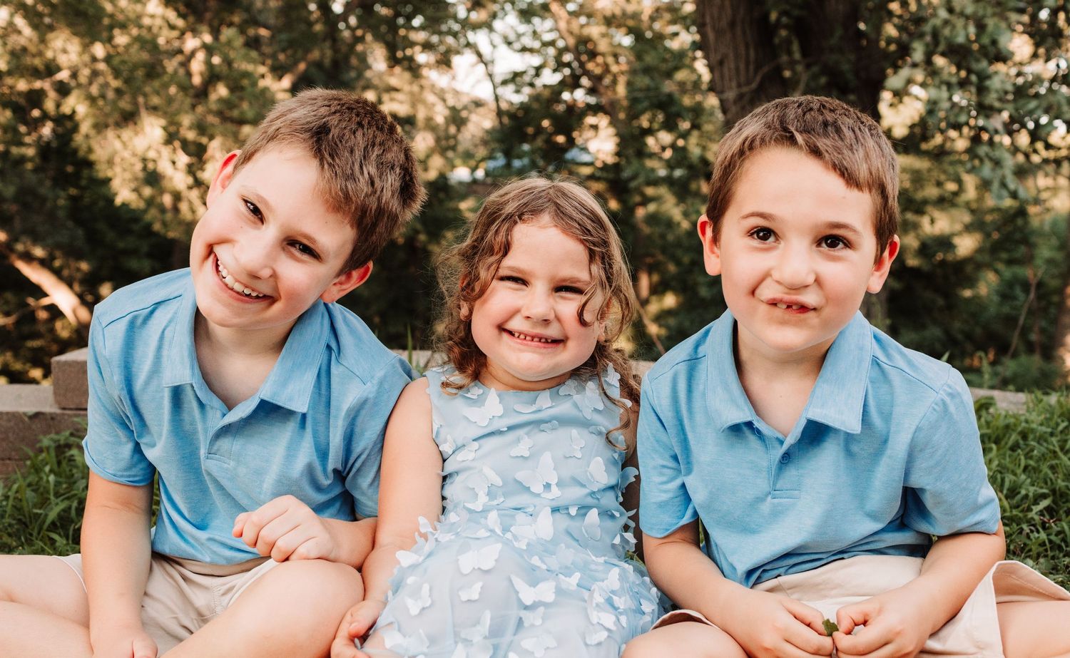 Three siblings in matching blue outfits sitting together smiling outdoors.