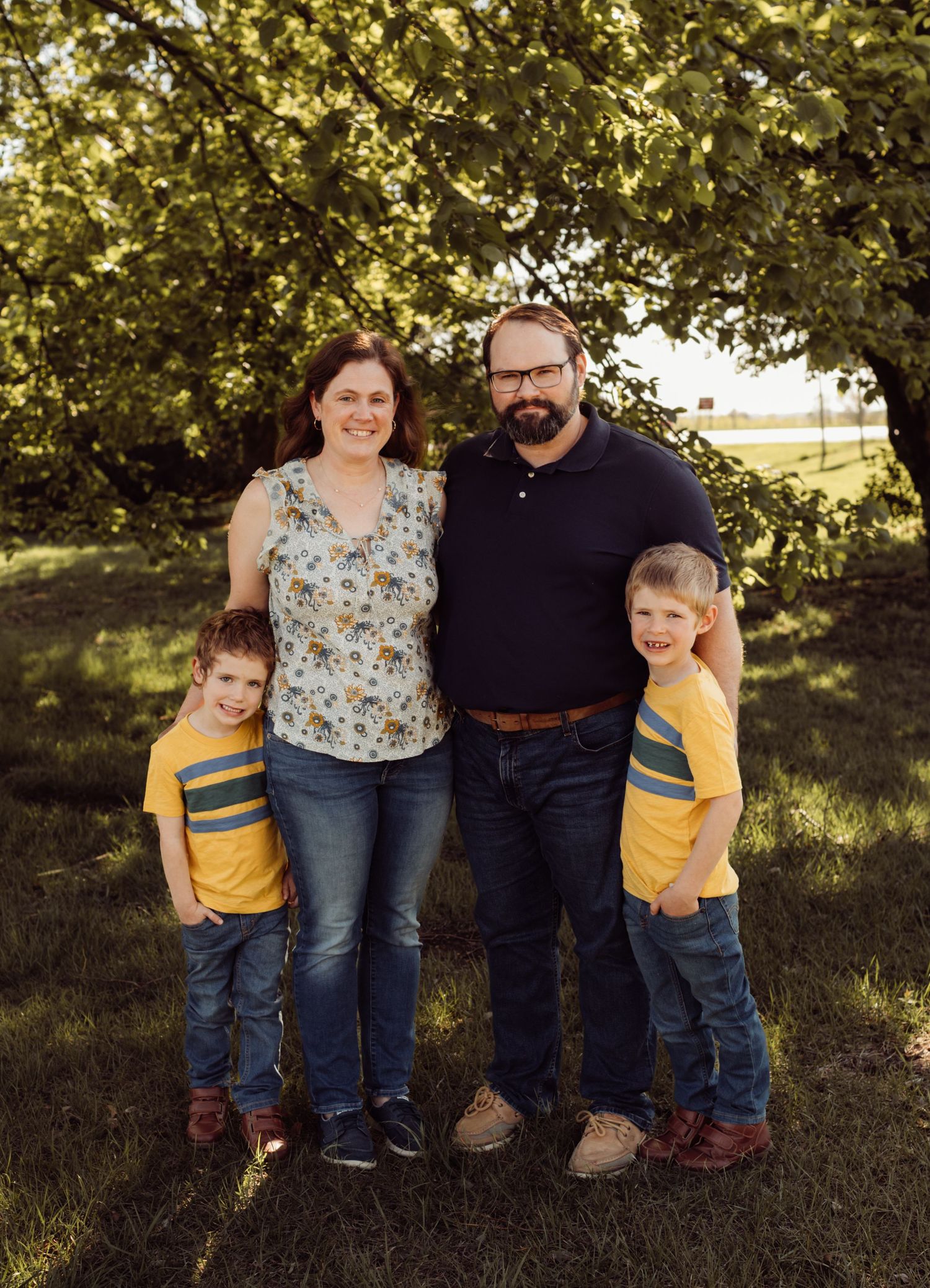 Family of four with two young boys in yellow shirts posing outdoors under a green tree in golden light.