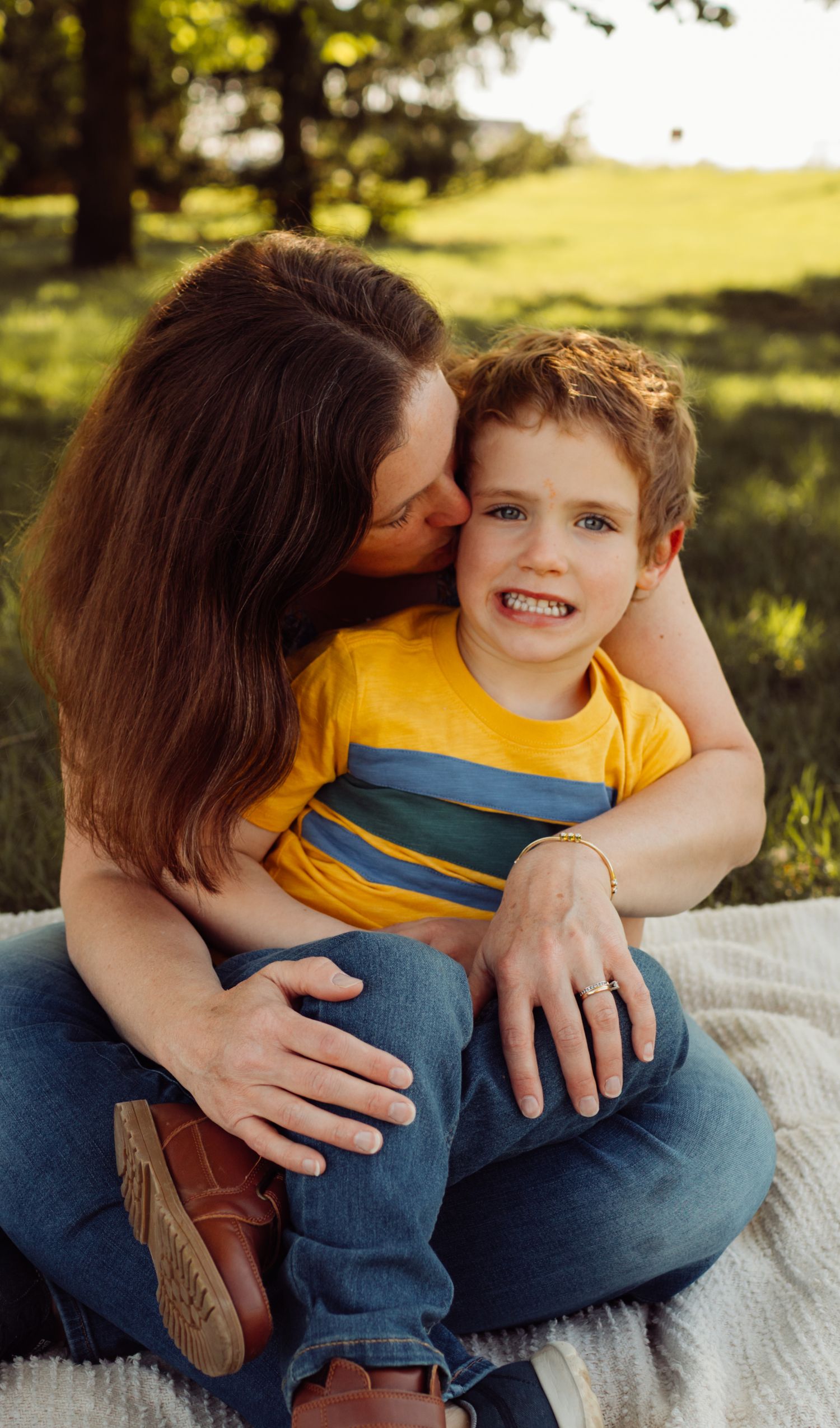 Mother kissing her smiling young son on the cheek while sitting on a blanket in a sunny green field.