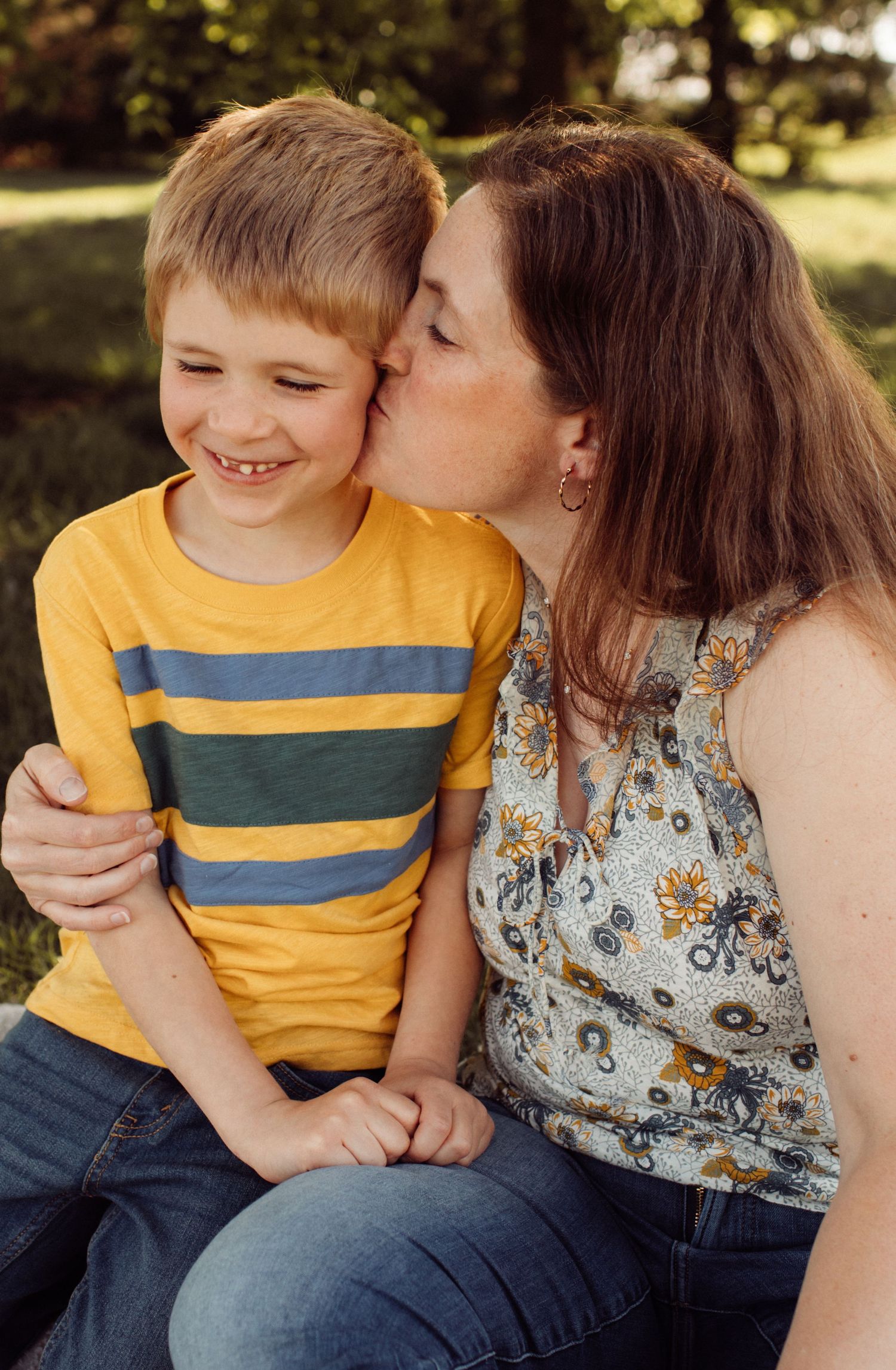 A mother kisses her smiling young boy on the cheek outdoors in a sunny park setting.