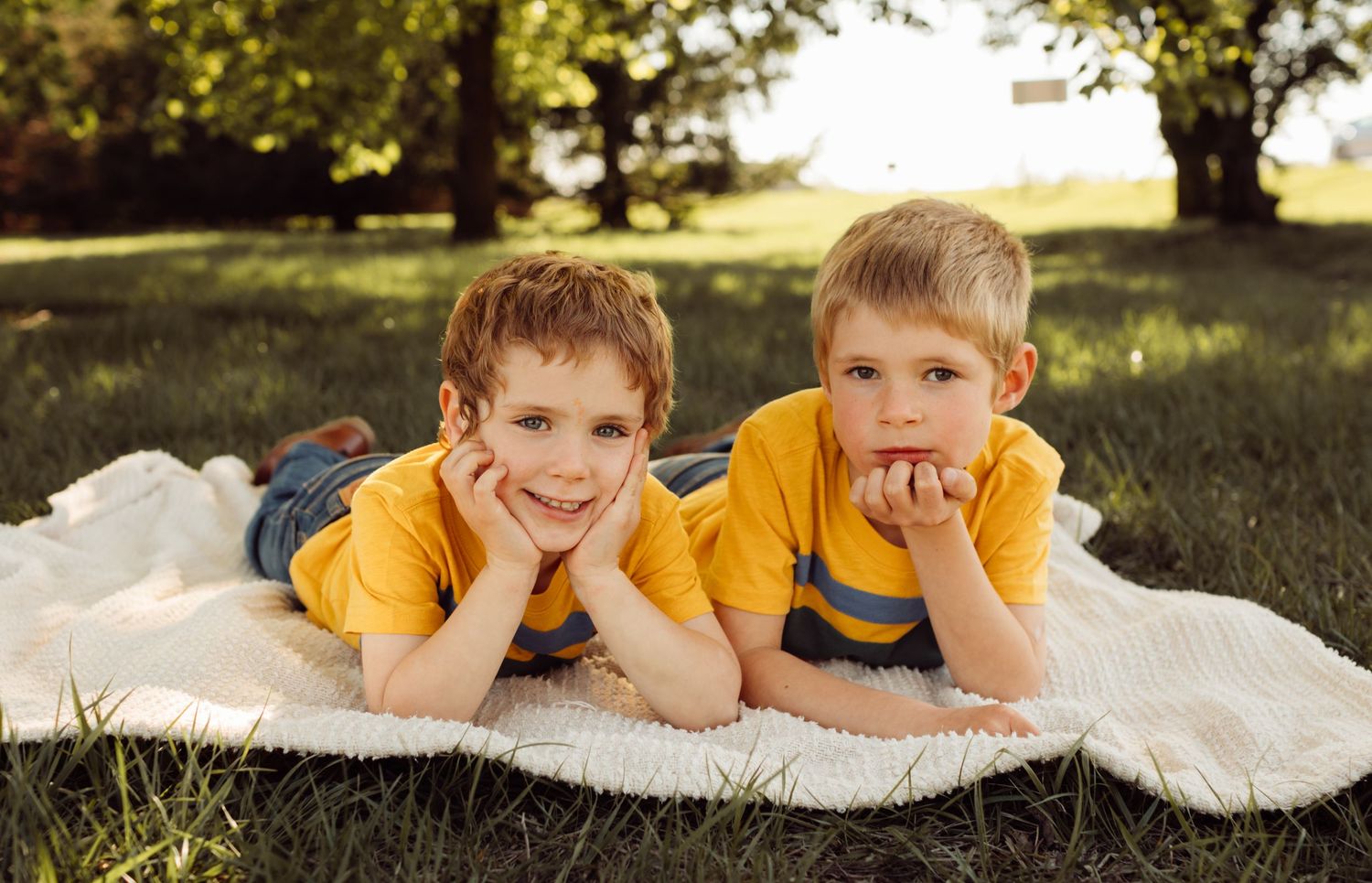 Two young boys in matching yellow shirts lie on a blanket in green grass smiling.