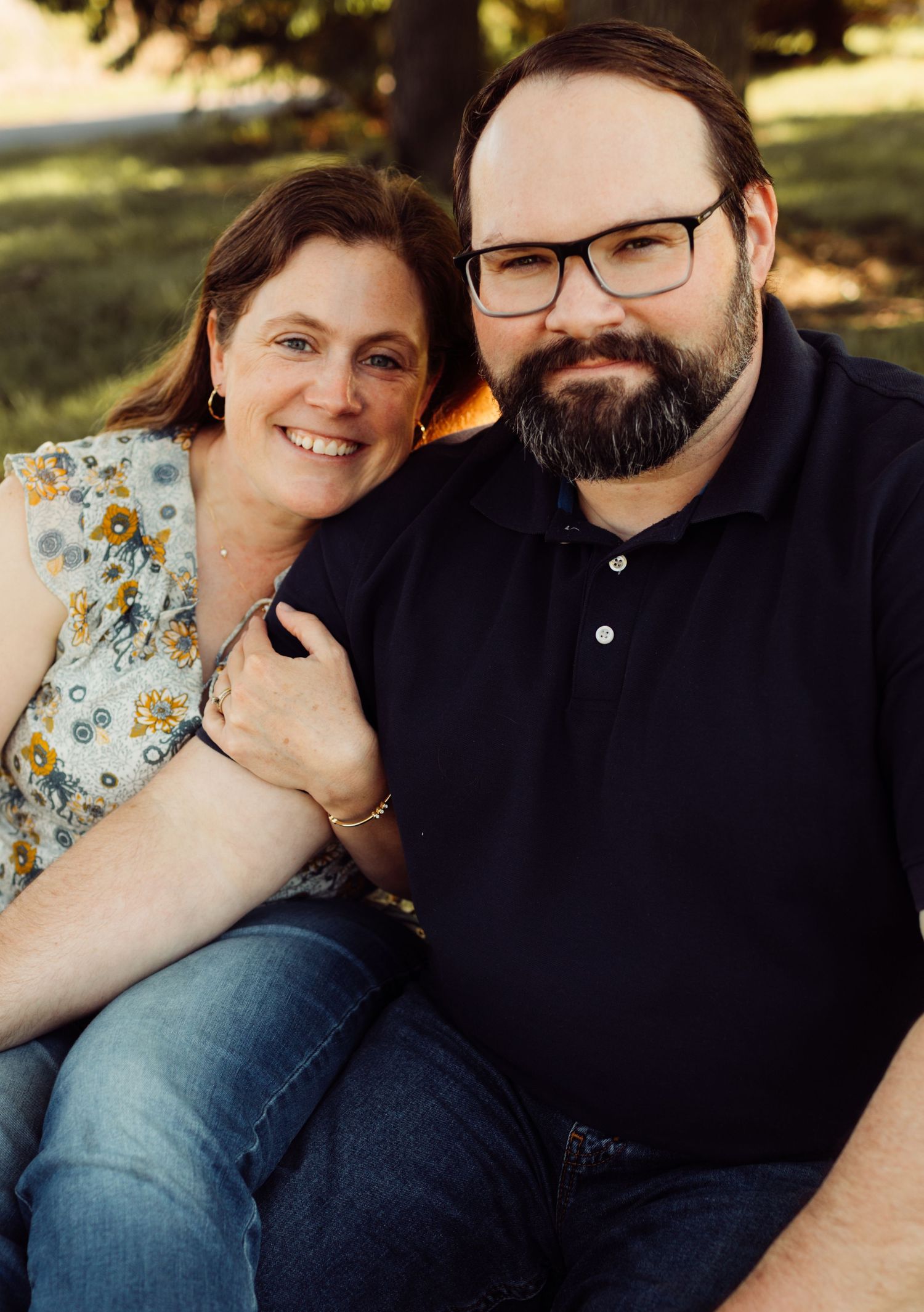 A smiling couple poses together outdoors, woman in floral top and man in navy polo shirt.