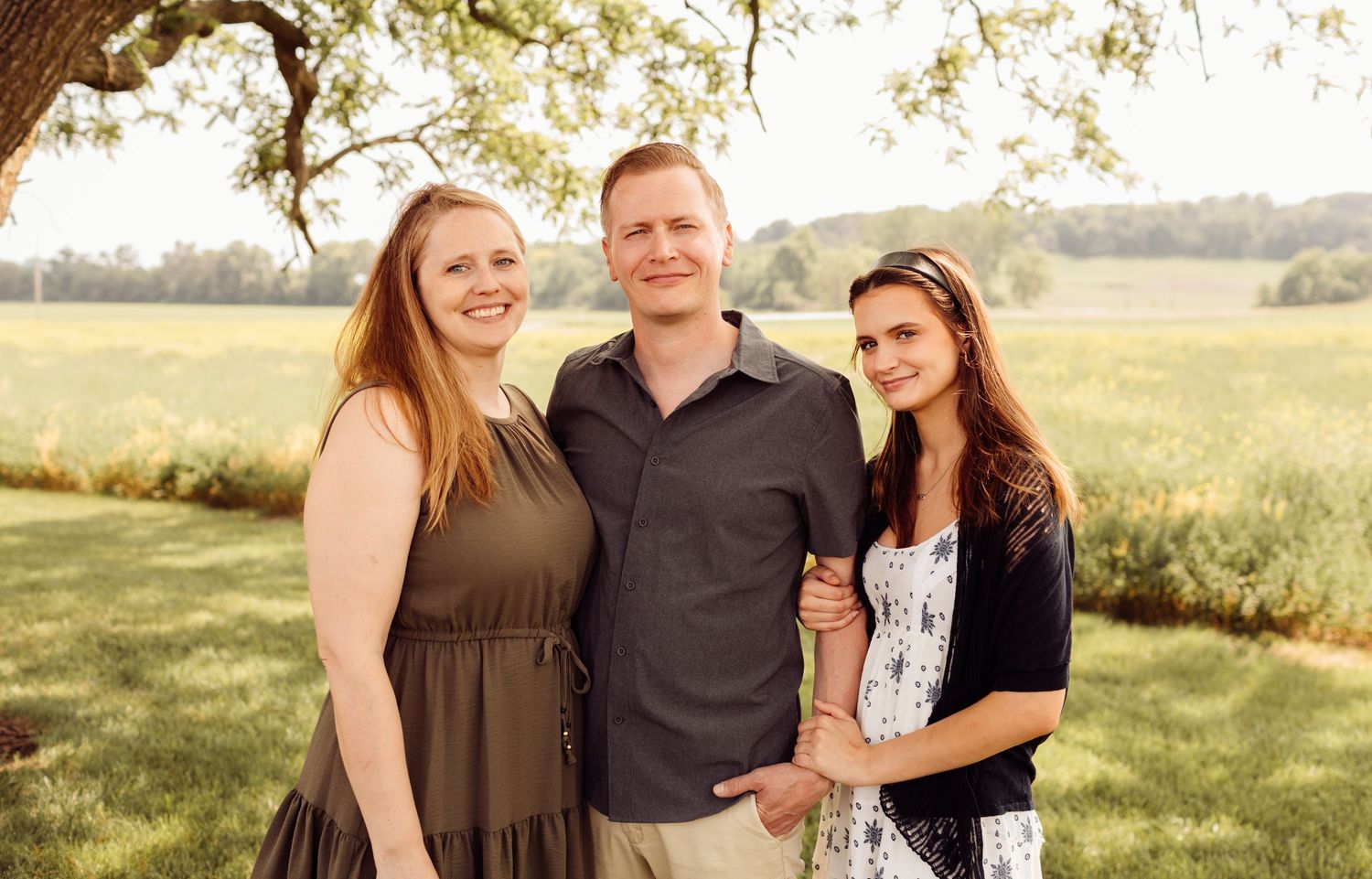 Man posing with two women outdoors under a tree in a green field during golden hour.
