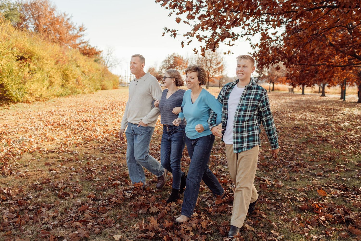 Family of four walking through an autumn orchard with colorful fallen leaves on the ground.