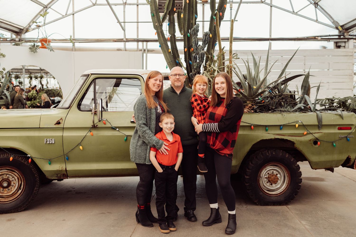 Family of five posing together in front of a vintage green pickup truck decorated with Christmas lights.