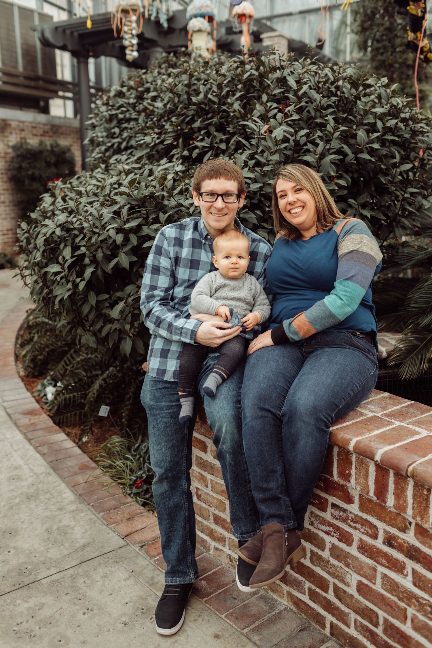 Young family with baby sitting on brick wall surrounded by lush greenery for a portrait photo.