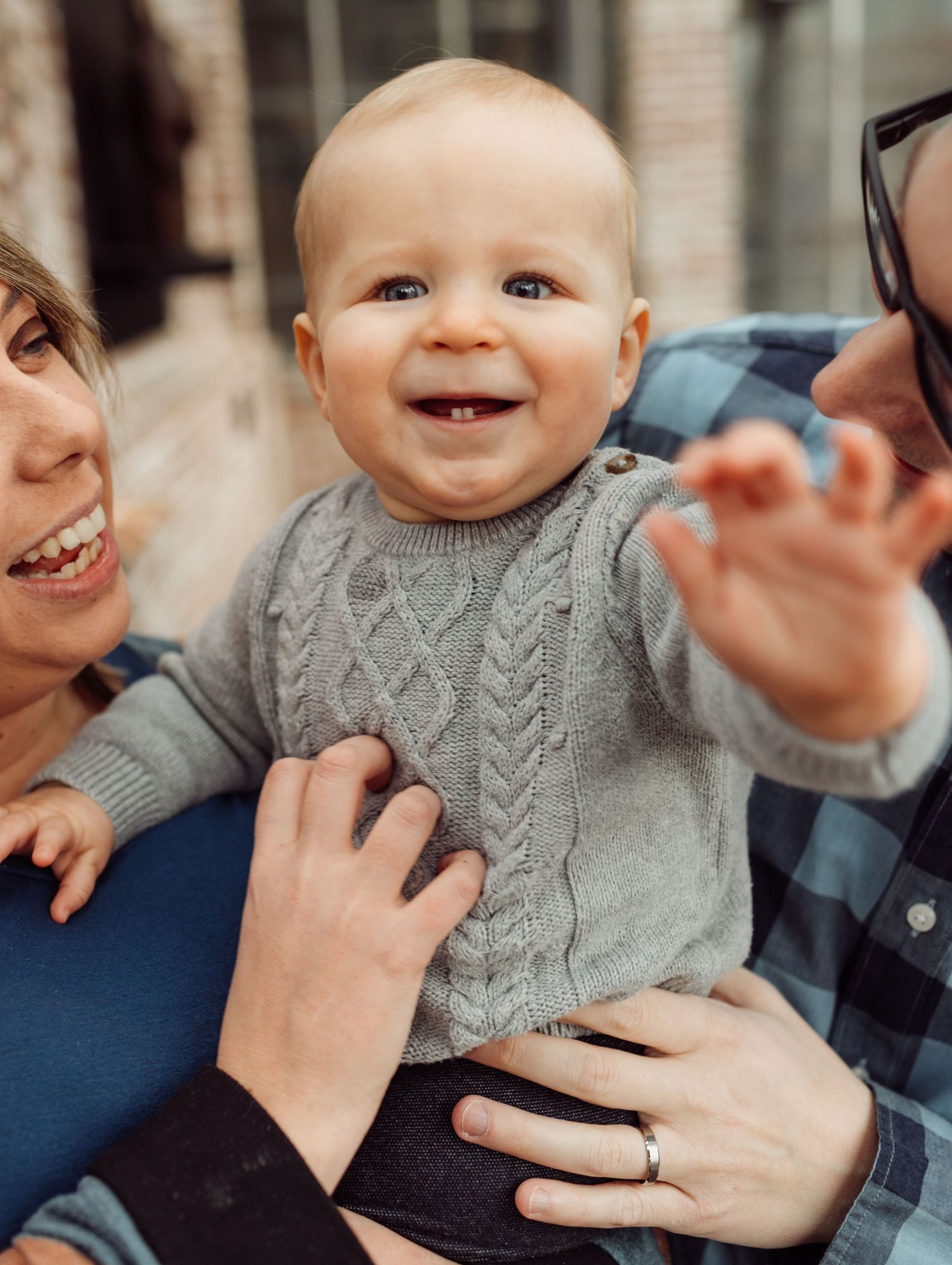 Happy baby in gray knit sweater smiling and waving while being held by parents outdoors.