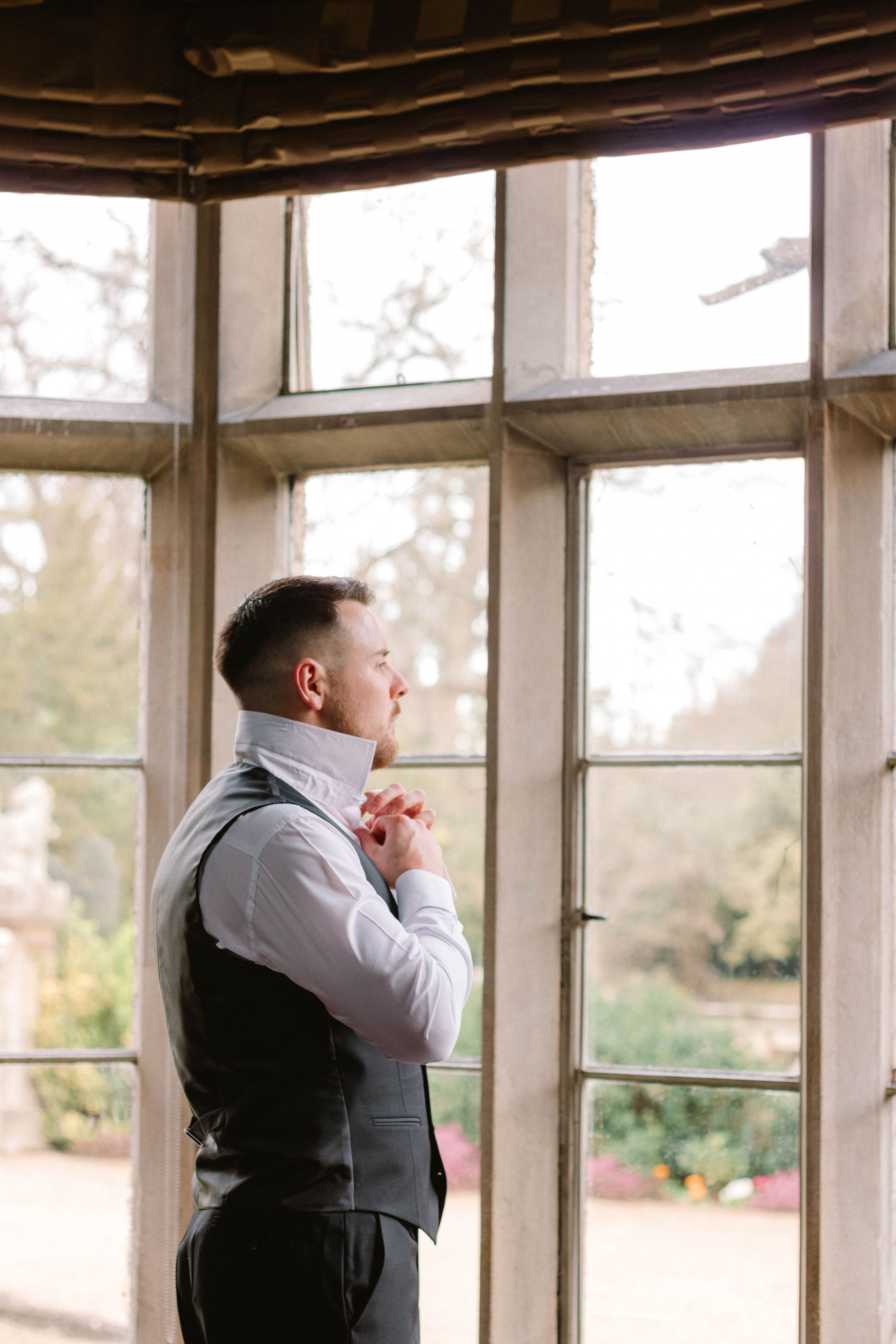 A person in formal attire stands thoughtfully by large windows in a sunlit room.