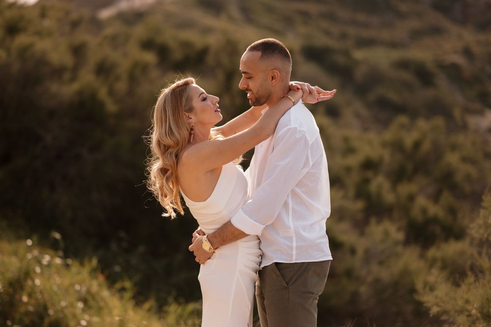 Couple embracing in golden sunset light outdoors, woman in white dress smiling at man in white shirt.