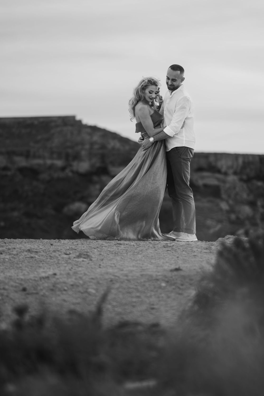 Black and white photo of a couple embracing outdoors, woman in flowing dress, man in white shirt, rocky landscape background.