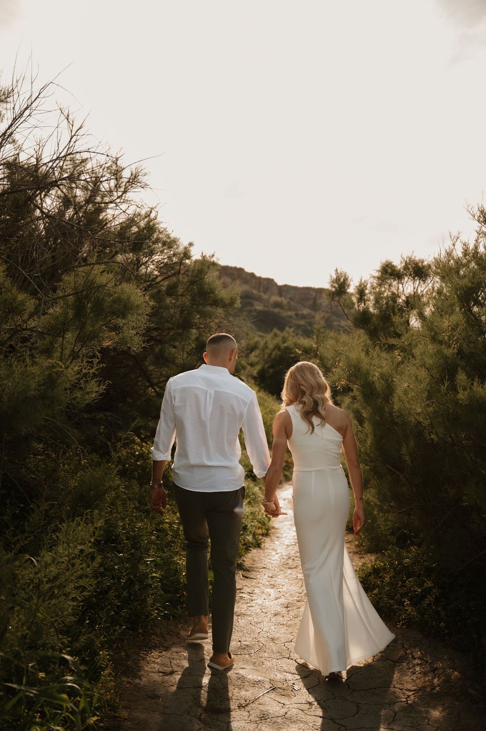 Couple holding hands walking down a scenic stone path surrounded by lush greenery at golden hour.