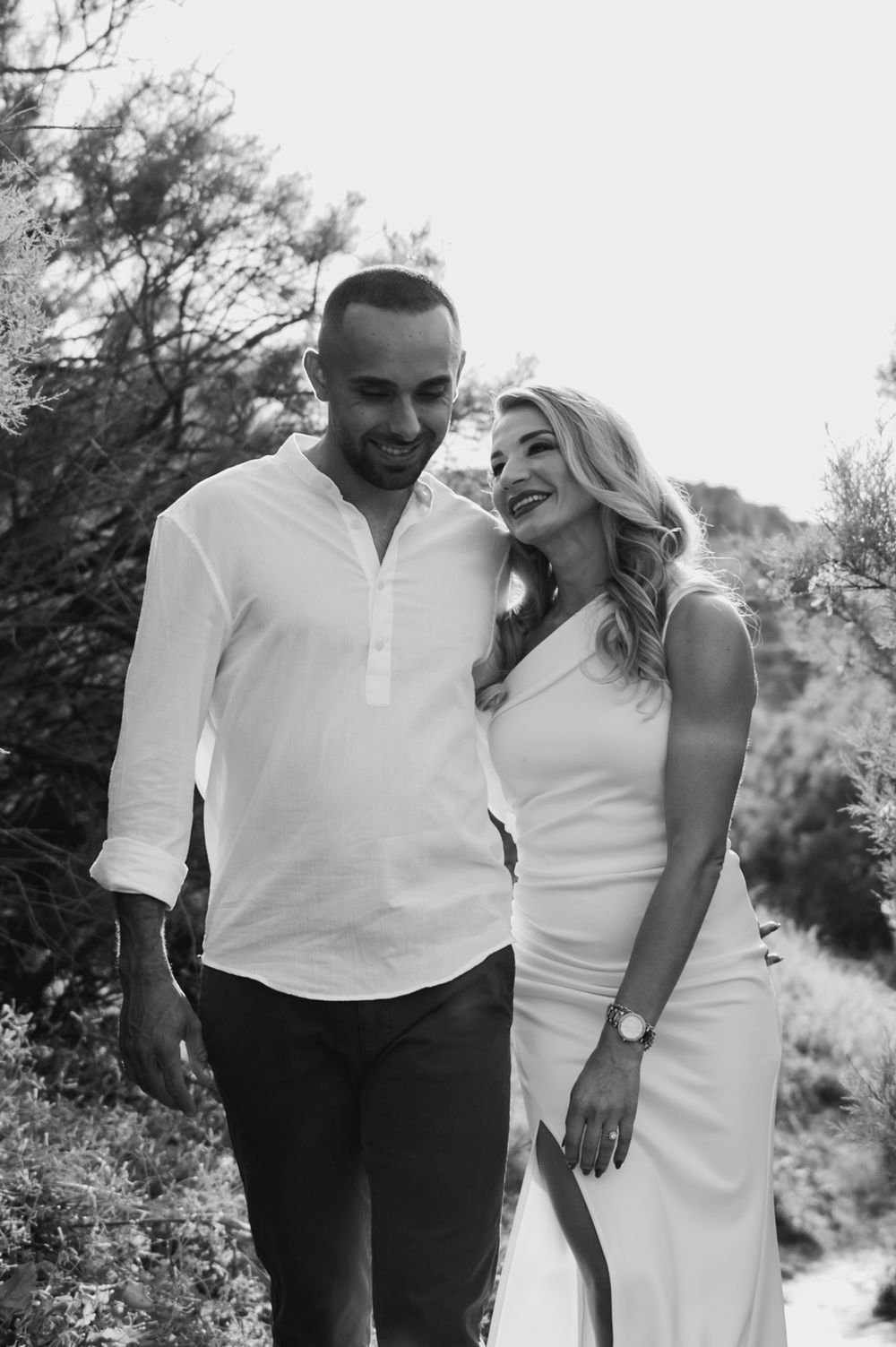 Black and white photo of a smiling couple in white outfits walking outdoors among trees.
