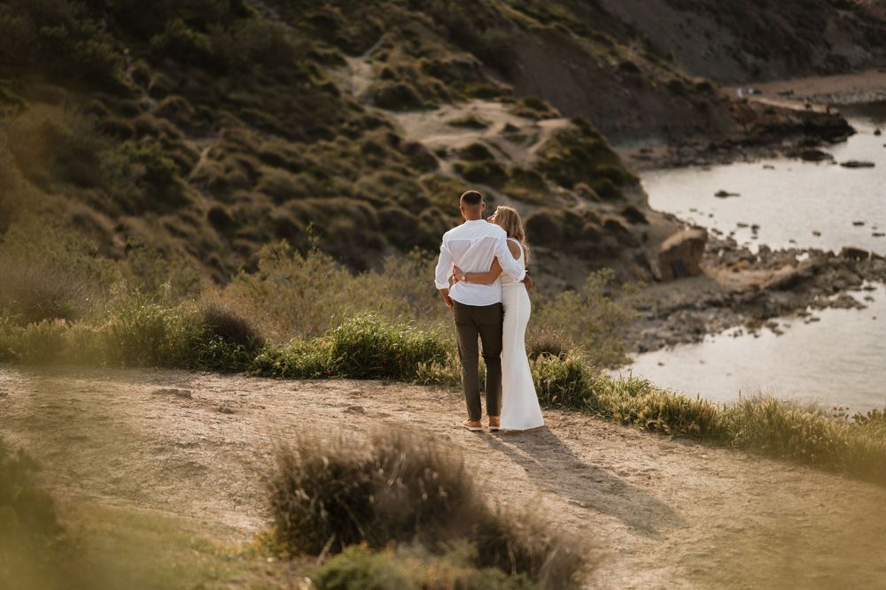 Couple in wedding attire standing together on a cliff overlooking a scenic coastal bay at golden hour.