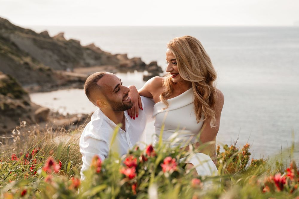 Couple sitting in a flower field by the ocean, sharing a romantic moment with rocky cliffs in the background.