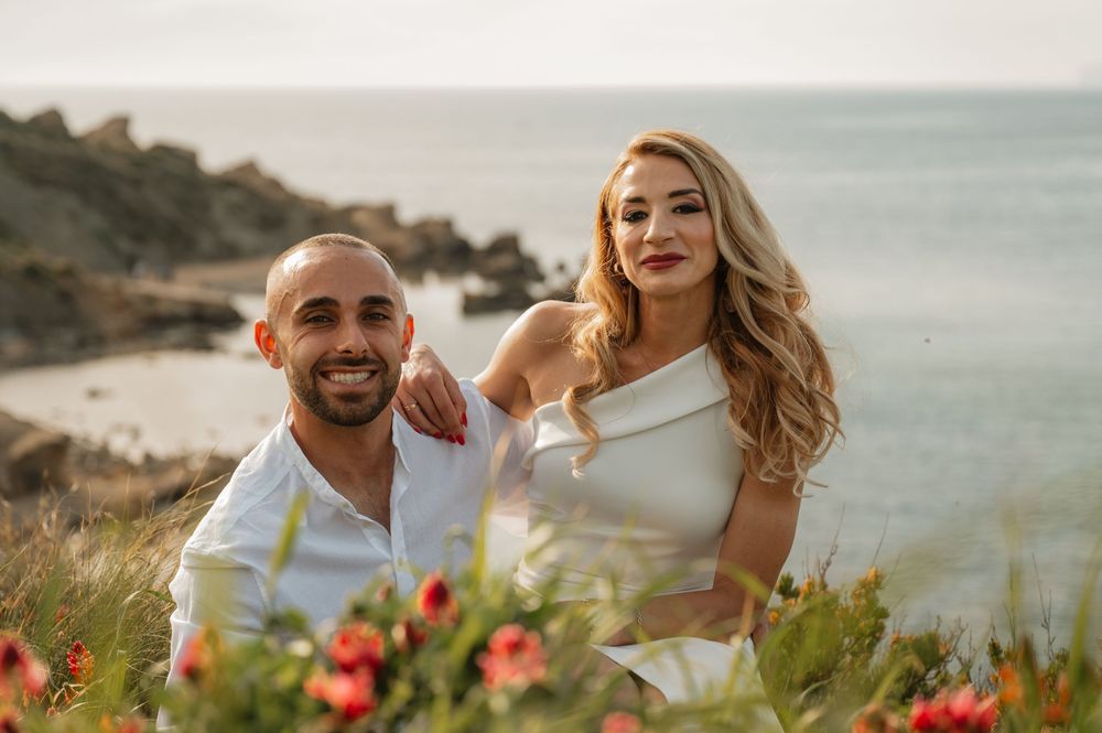 Couple posing together outdoors near rocky coastline, woman in white dress with blonde hair, man in white shirt smiling.