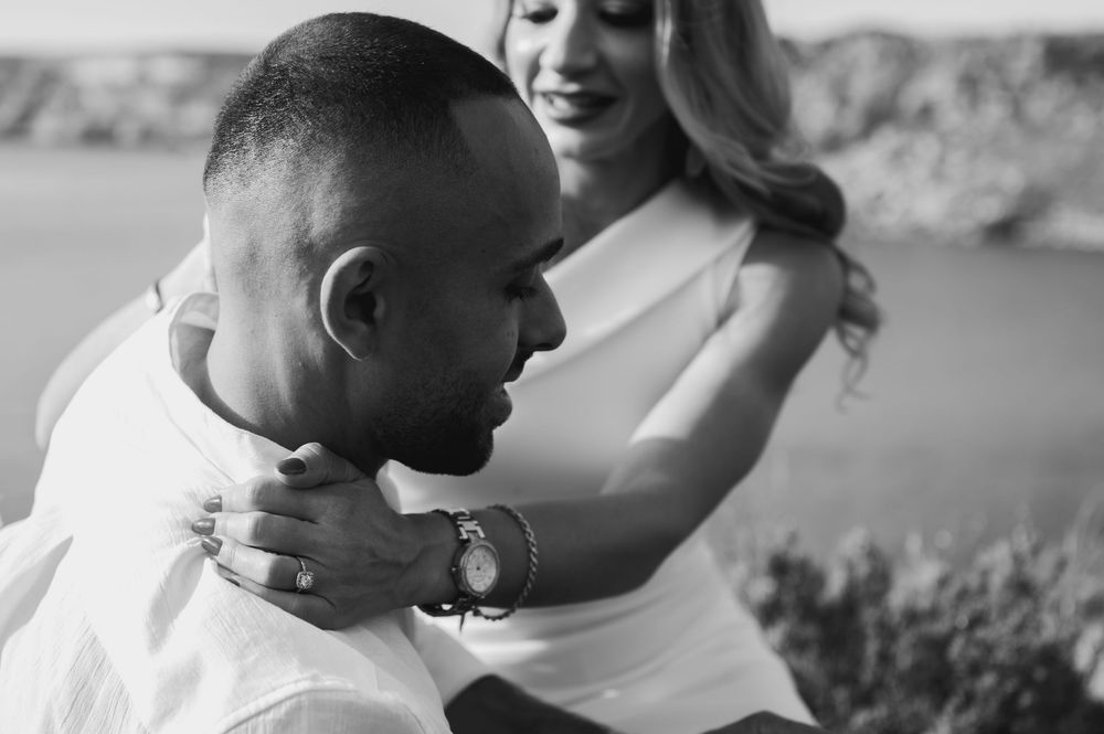 Black and white photo of a couple embracing outdoors, woman in white dress placing hands on mans shoulders.