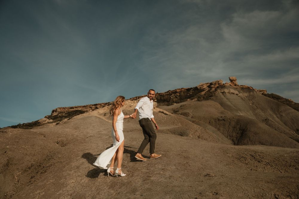 Couple holding hands on rocky desert hillside, woman in white slit dress, dramatic sky backdrop.