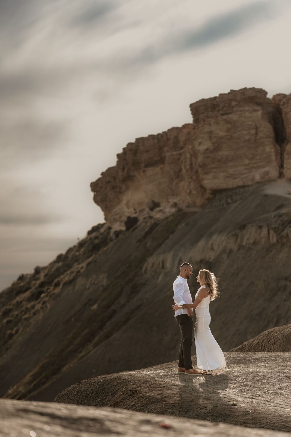 Couple in wedding attire standing on rocky terrain with dramatic sandstone cliffs under cloudy sky.