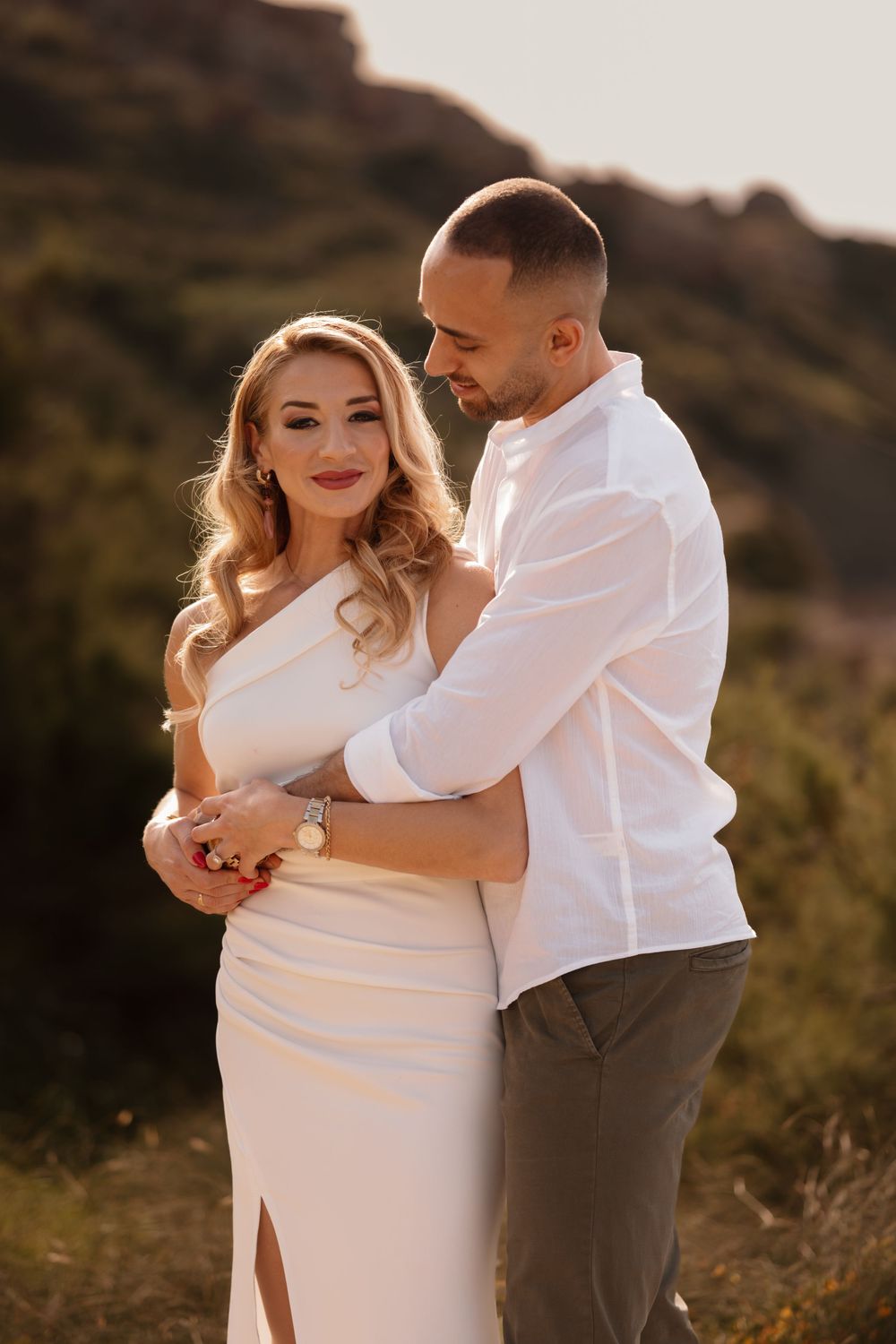 Couple embracing at golden hour, woman in white dress, man in white shirt, with green hills in background.