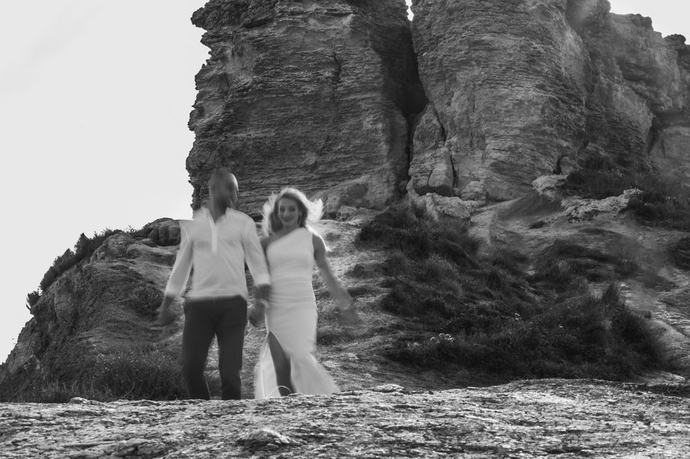 A couple in wedding attire holds hands on a rocky beach in a romantic black and white photo.