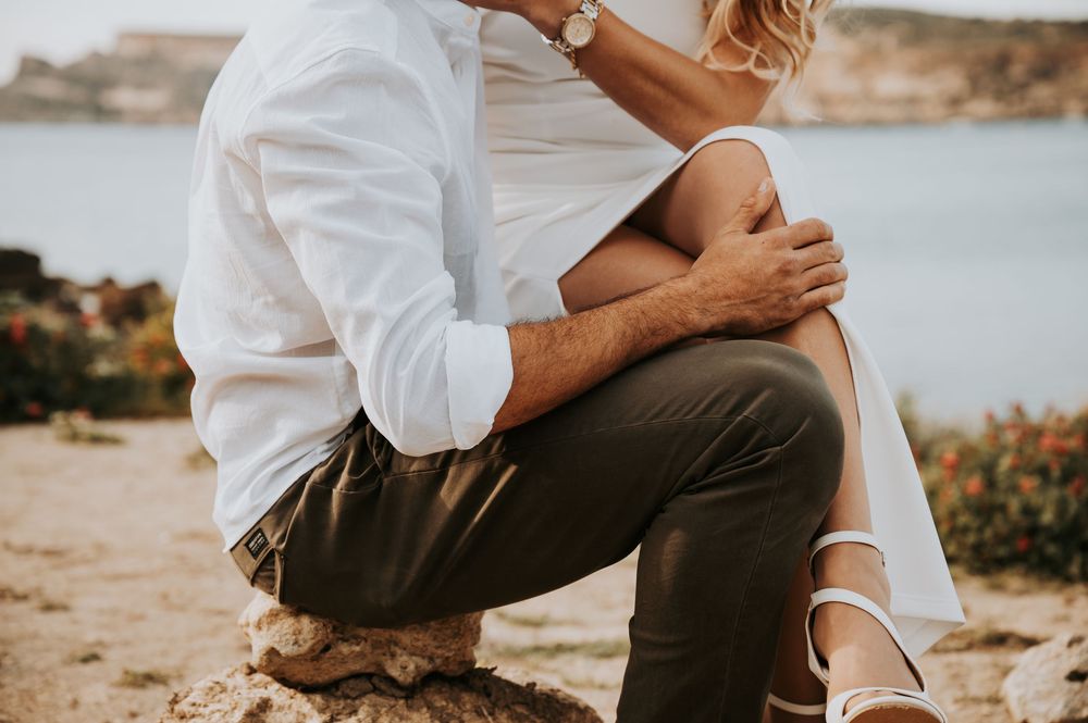 Close-up of couple embracing on rocky shore, man in white shirt and olive pants, woman in white dress.