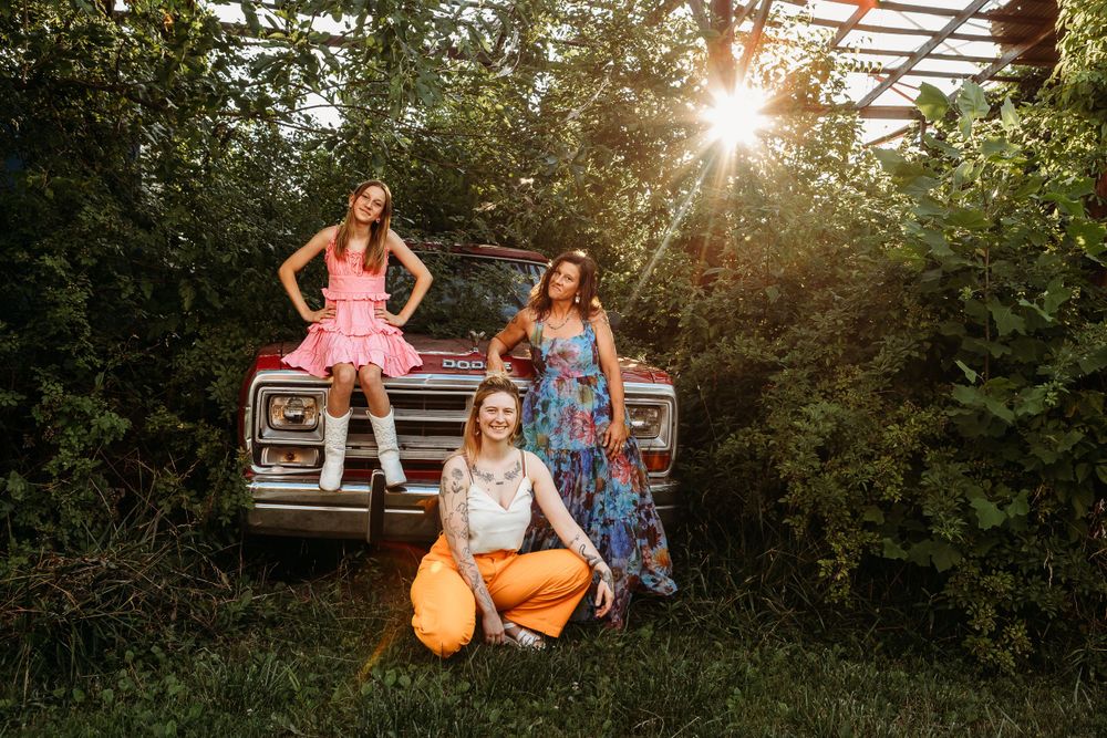 Three friends pose on a vintage truck during golden hour in an overgrown garden.
