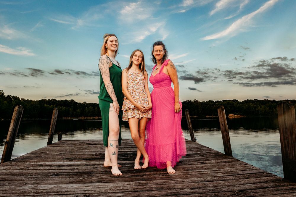 Three friends in colorful dresses pose on a dock at sunset.