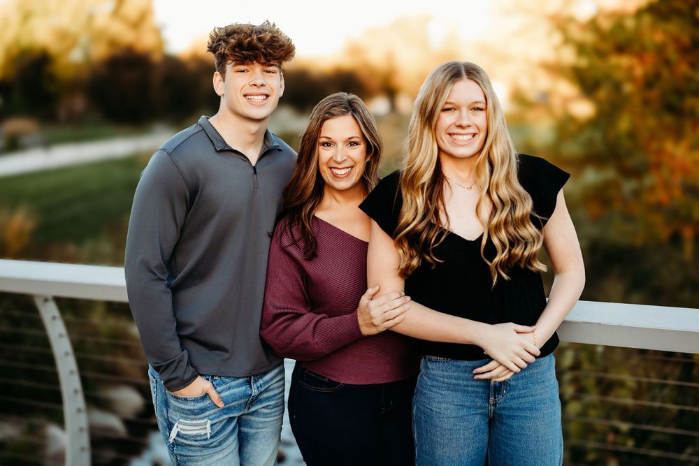 Three people in casual fall attire pose together on a bridge during sunset.