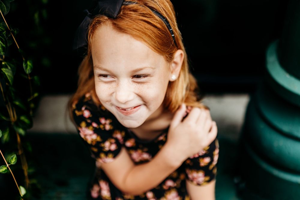 Child with red hair smiles while wearing floral dress outdoors.