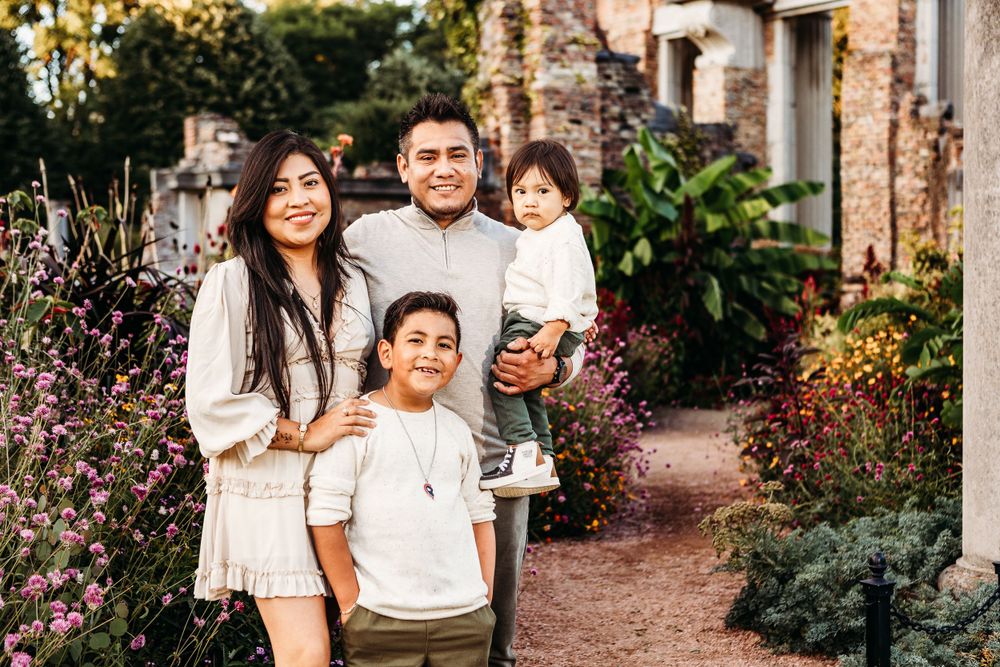 A family of four poses together in light-colored outfits near stone ruins and purple flowers.