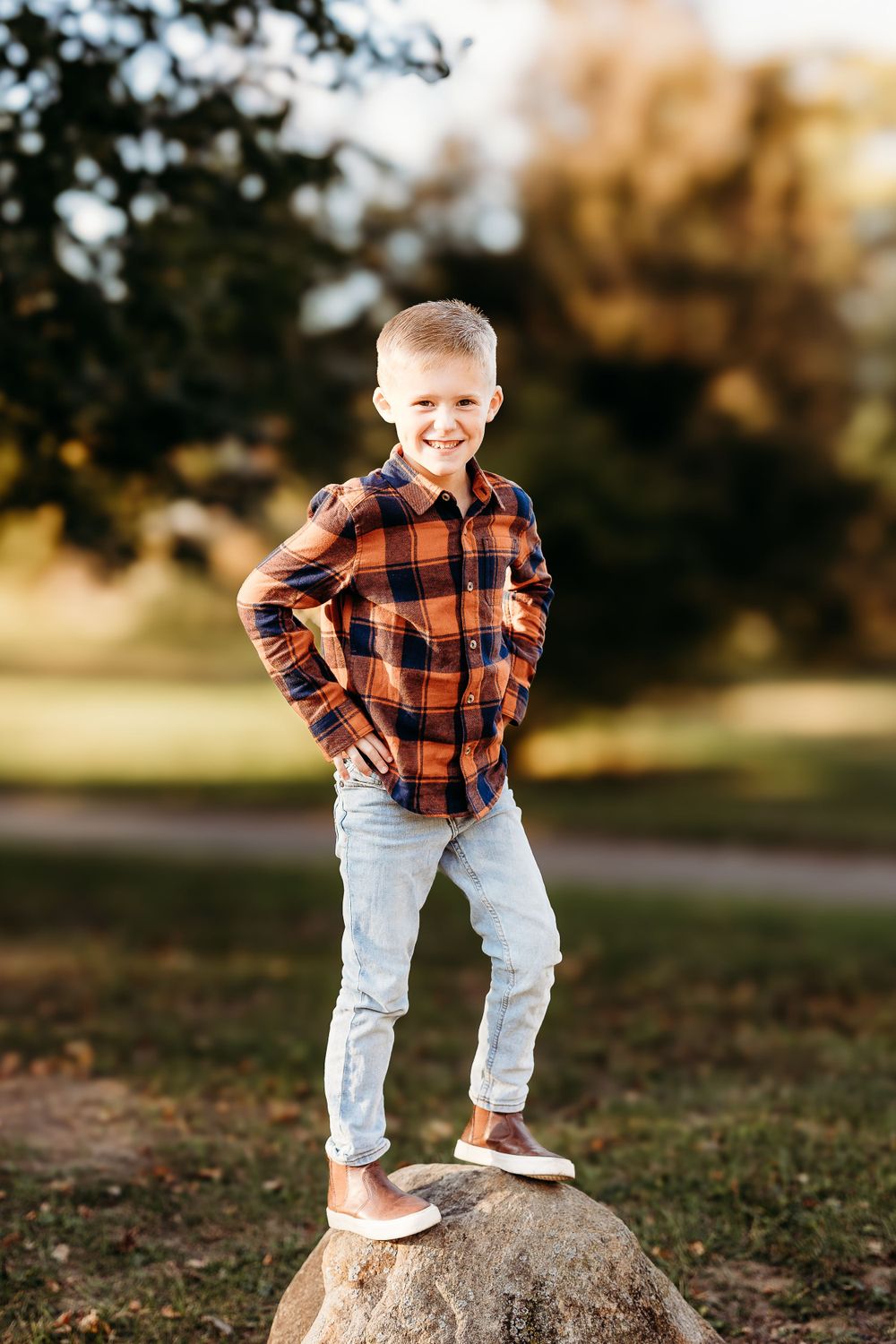 Child in plaid shirt stands confidently on rock in outdoor autumn setting.