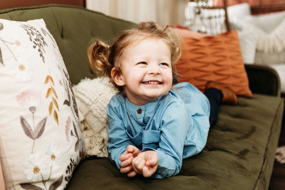 Toddler in blue outfit smiling brightly while sitting on olive green couch.