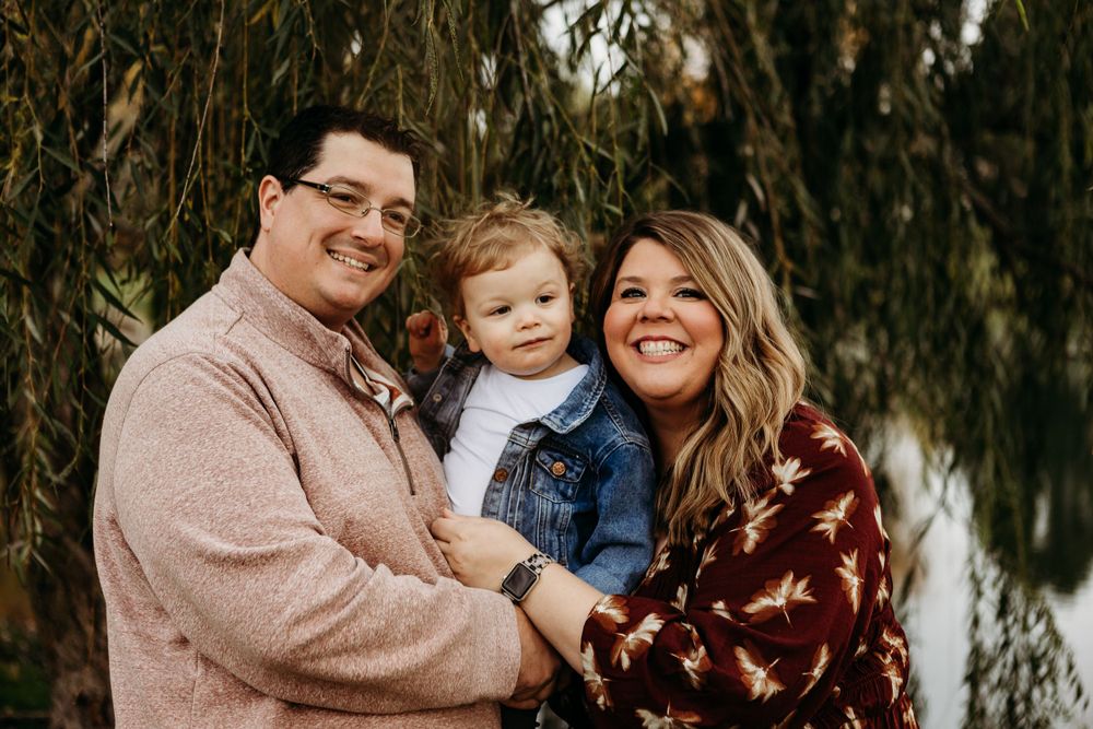 Family of three pose together under willow tree branches during fall photo session.