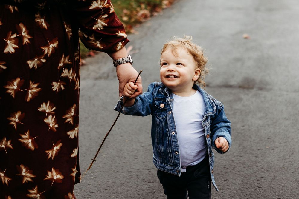 Toddler in denim jacket walks along path holding stick while smiling with joy.