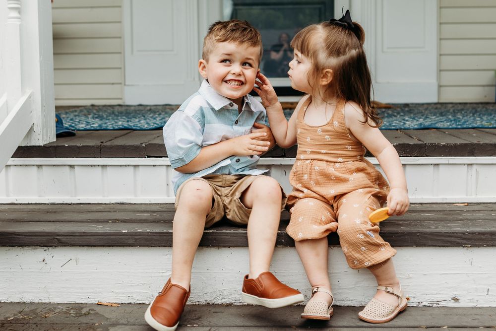 Two young children sit together on wooden steps wearing coordinated brown leather shoes.