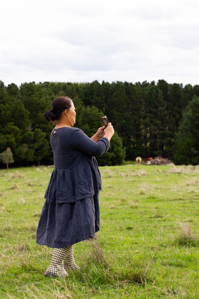 A woman in a navy linen dress and gumboots taking a photo in a green paddock with dark pine forest in the background. Image by Em Callaghan