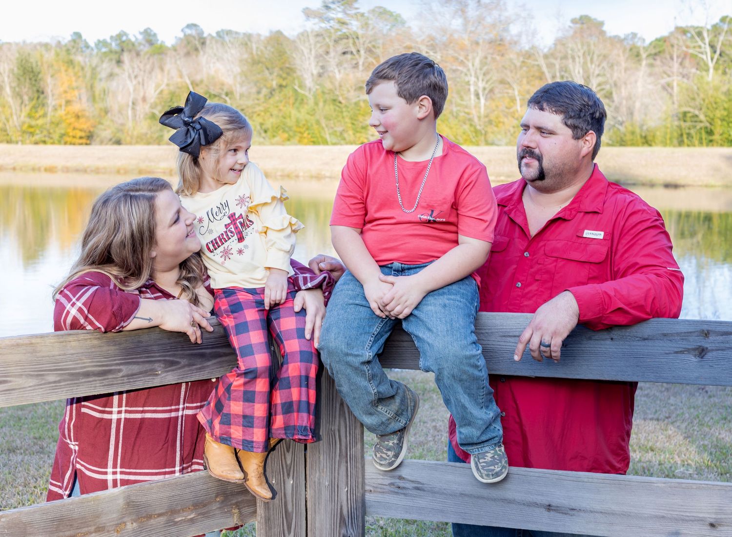Family in coordinating red outfits sitting together on wooden bench by lake during autumn afternoon.