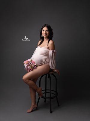 Smiling studio maternity portrait with pink flowers and a soft peach bodysuit, professional photography by Precious Pictures, Malta.
