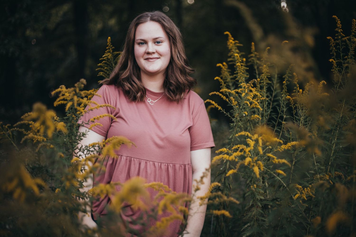 A person in a pink dress stands among tall yellow wildflowers in a dark nature setting at dusk.