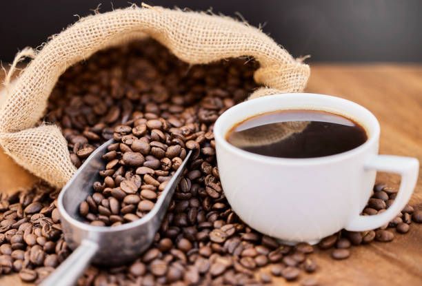 A white coffee cup with fresh brewed coffee sits beside scattered roasted coffee beans and a burlap sack.