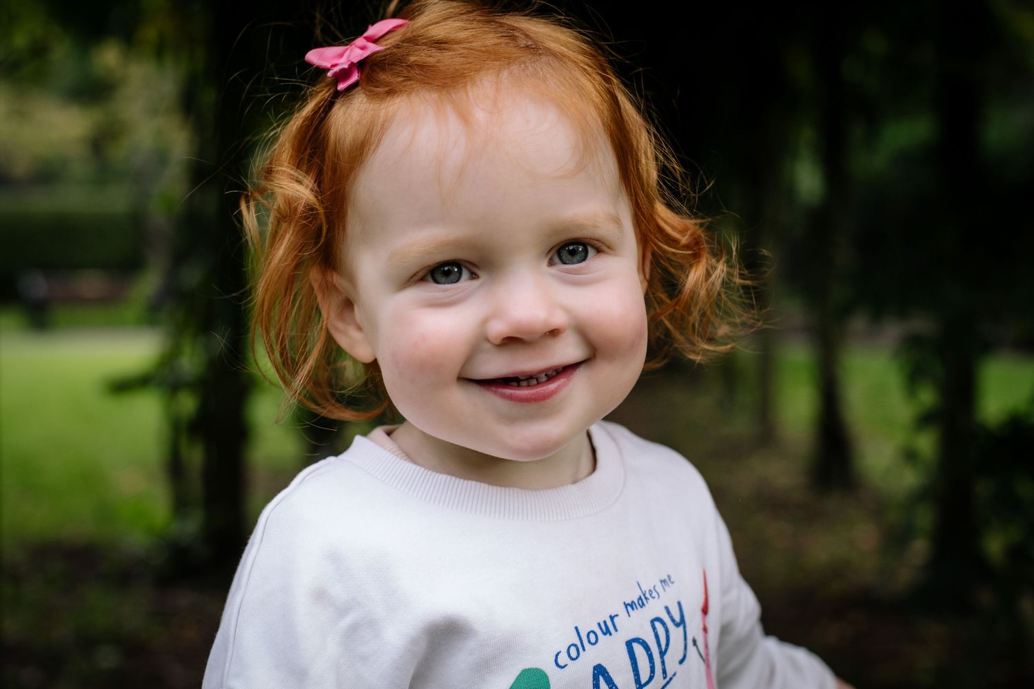 A cheerful child with red hair wearing a white shirt smiles outdoors in a green park setting.