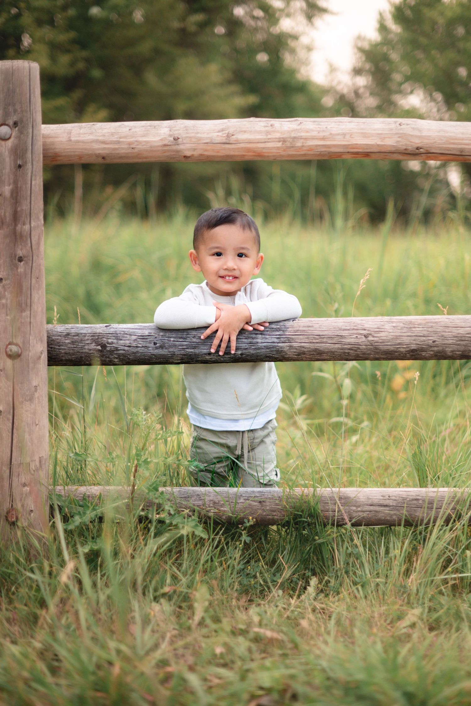 Small child stands behind rustic wooden fence in a grassy field during golden hour.