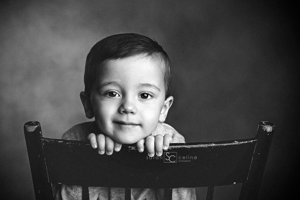 Dramatic black and white portrait of child resting chin on wooden chair back.