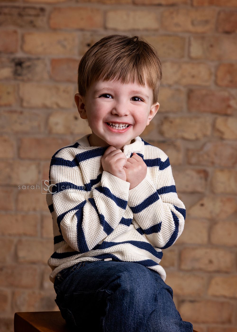Child in striped sweater poses playfully on wooden stool against brick wall.
