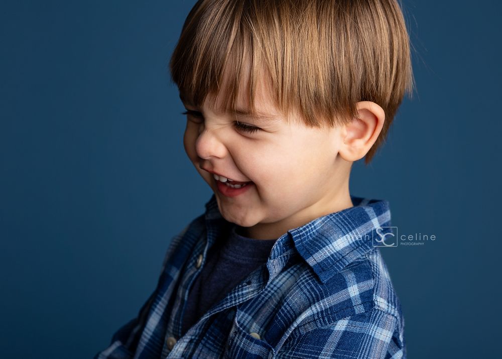 A young child in a blue plaid shirt smiles while looking off to the side against a dark blue background.