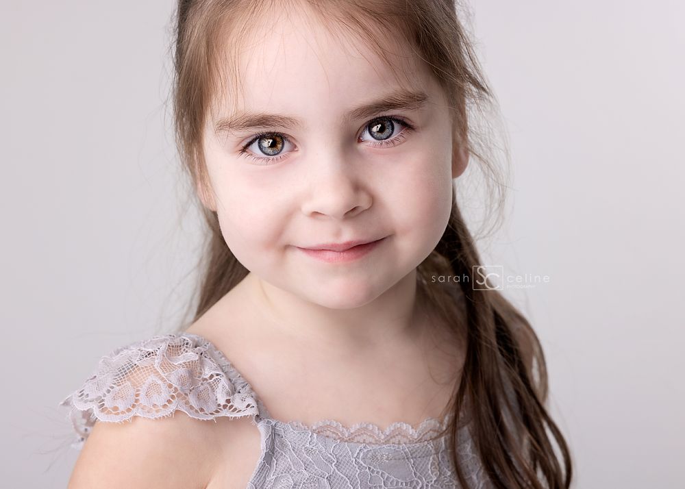 Studio portrait of young girl in lace dress with soft smile against light background.