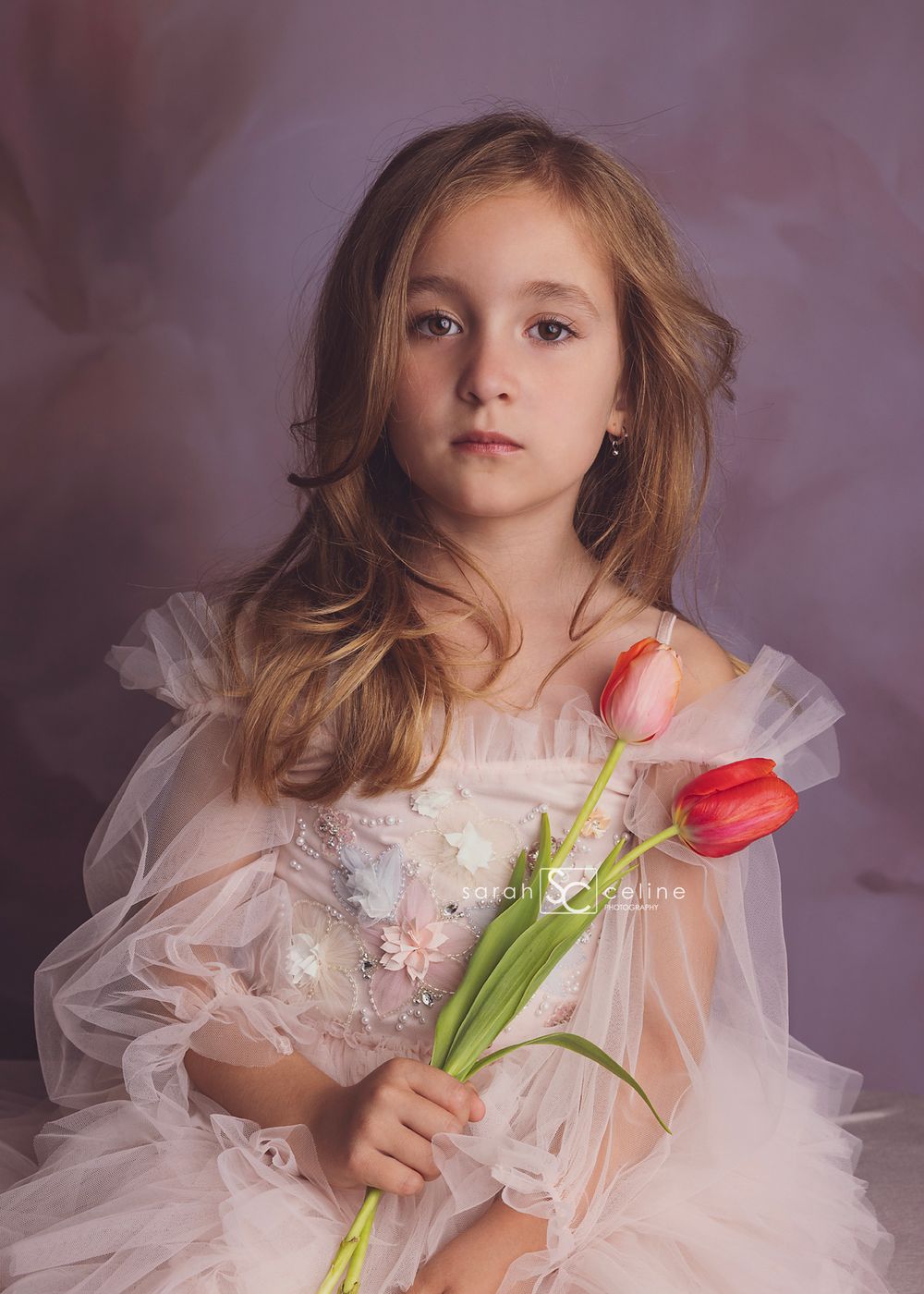 Young child in soft pink tulle dress holding two fresh tulips in ethereal portrait setting against mauve background.