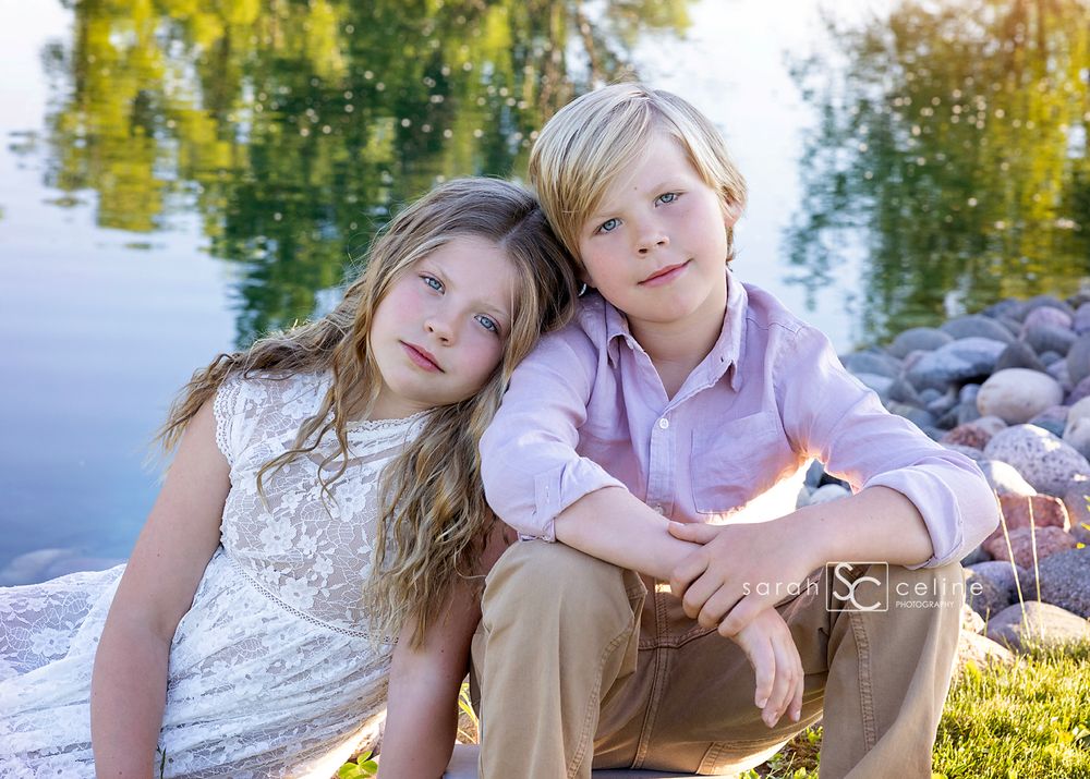 Two children sit casually by a serene lake with autumn foliage reflected in the water.